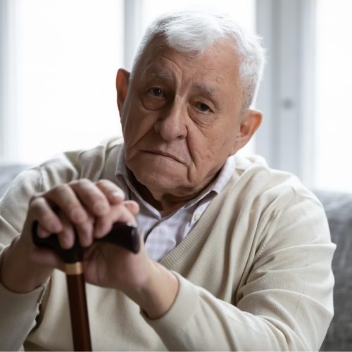 Head shot portrait depressed lonely elderly man with wooden cane looking at camera, frustrated mature male folded hands on walking stick, sitting on couch alone, loneliness and solitude