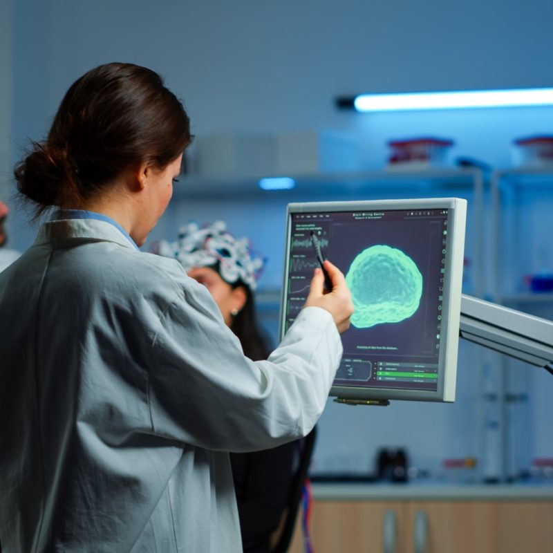 Researcher looking at monitor analysing brain scan while coworker discussing with patient in background about side effects, mind functions, nervous system, tomography scan working in laboratory