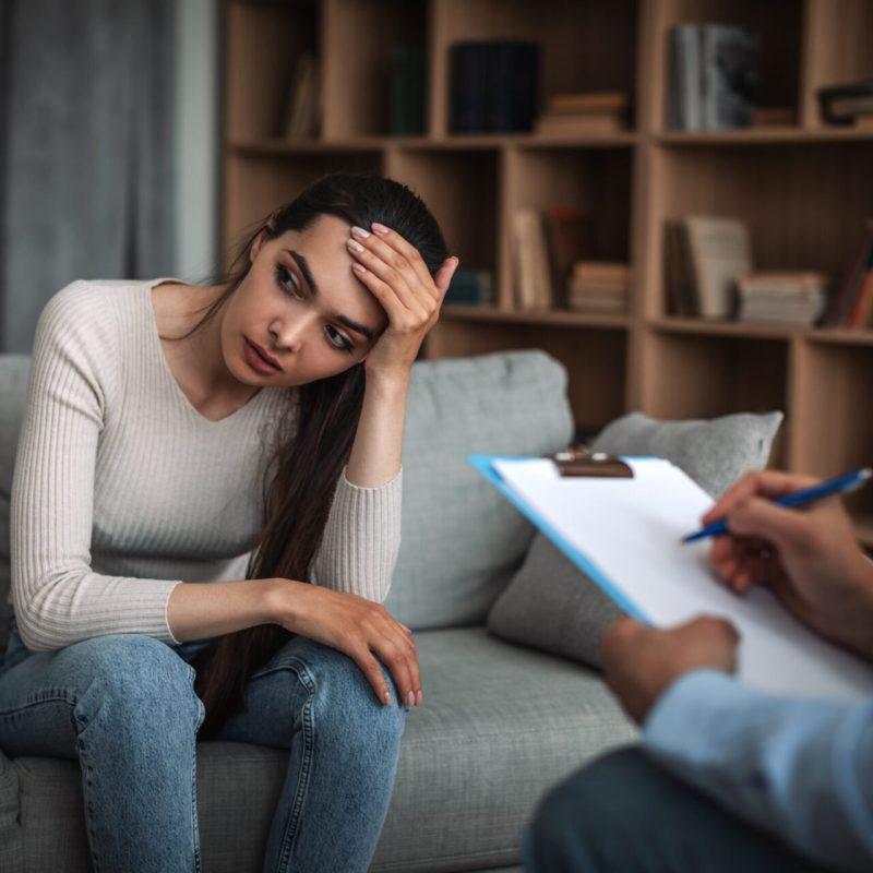 Sad young european lady hold her head with hand at consultation with male doctor psychologist with tablet in clinic interior. Medical professional support, help and health care, psychological problems